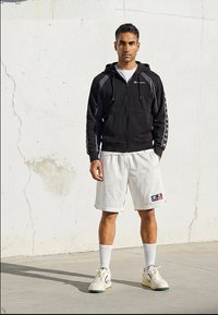 Mann står mot en hvit vegg iført en svart hettejakke med glidelås, hvite treningsshorts, hvite sokker og hvite joggesko.