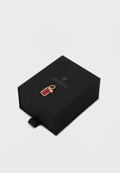 Sort boks med tekstureret overflade, der har en guldfarvet charm formet som en tag med en rød indsats og en lille bladdetalje samt en træksnøre.