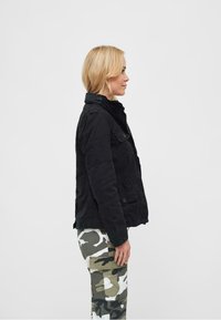 Giacca nera in stile militare con dettagli in bottone, tasche laterali e colletto alto, indossata sopra pantaloni cargo mimetici con vari modelli in verde e bianco.