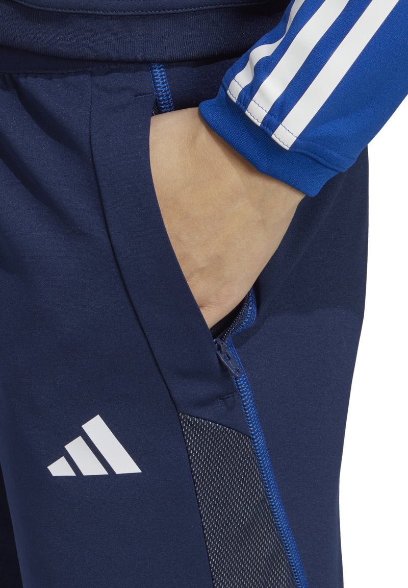 Pantalons de sport bleu marine avec une poche zippée, présentant un panneau en maille et le logo blanc d'Adidas. Accents bleus sur les poignets avec trois bandes blanches.