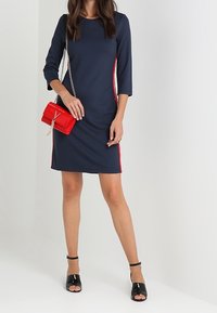 Abito blu con maniche a tre quarti e strisce rosse ai lati, abbinato a una borsa rossa e sandali aperti neri con tacco. Tessuto liscio, design aderente.