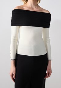 Off-the-shoulder top with black and white color block design, ribbed texture, and button accents at the cuffs. Slim fit and long sleeves.