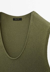 Olive green knitted tank top with a deep V-neck design. Features a textured fabric and a black label at the neckline.