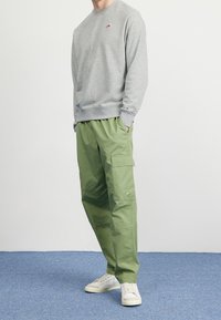 Grå sweatshirt med liten logotyp, gröna cargo-byxor med sidofickor och vita sneakers på en blå matta.