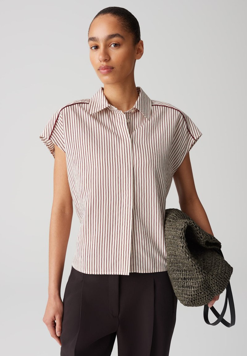 Camicia a maniche corte con colletto, a righe rosse e bianche sottili, realizzata in tessuto leggero. Il modello tiene una borsa scura e texturizzata.