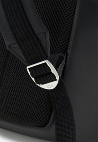 Svart ryggsekk med detaljer som en sølvspenne og teksturert mesh-reim. Materialet har en glatt overflate med et slitesterkt, strukturert design.