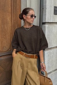 Brauner Strickpullover mit kurzen Ärmeln, beigefarbene, plissierte Hose und ein beiger Gürtel. Die Accessoires umfassen eine Perlenkette und eine braune Handtasche.