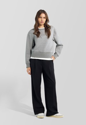 Joven mujer con el pelo largo y castaño que lleva una sudadera gris, pantalones anchos negros y zapatillas blancas de pie delante de un fondo claro y liso.