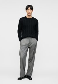 Homme portant un pull noir à manches longues avec des rayures texturées, un pantalon gris plissé et des chaussures en cuir noires, debout les mains dans les poches.