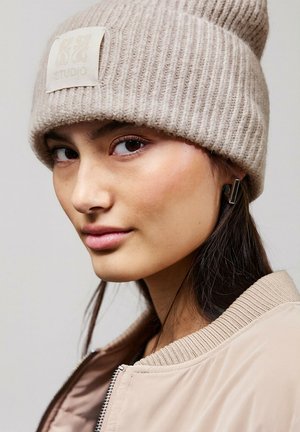 Jasnobeżowa prążkowana dzianinowa czapka z kwadratową metką z napisem "S&D STUDIO." Modelka nosi beżową kurtkę bombkę z teksturowanym kołnierzem.