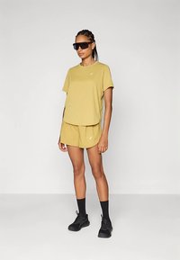 Mustársárga pamut póló és rövidnadrág szett; laza fazon, kis logó részlettel. Fekete sportcipővel és napszemüveggel párosítva.