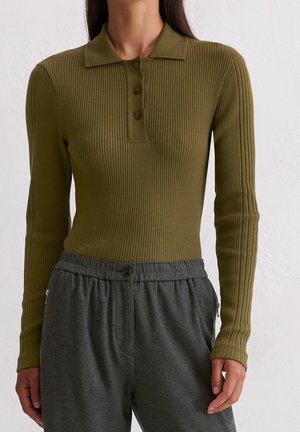 Kobieta w oliwkowozielonej, prążkowanej koszulce polo z długim rękawem, włożonej w wysokowalne ciemnoszare spodnie z zapinanym na guziki paskiem.