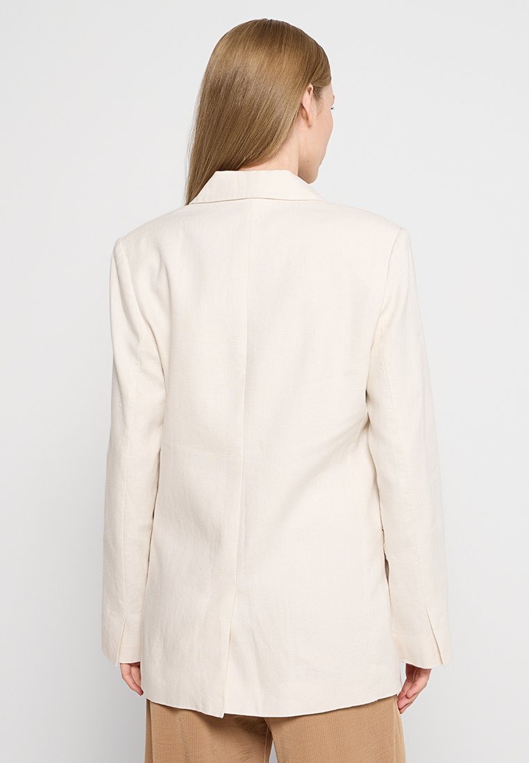 Blazer en lin beige clair avec revers, fente arrière unique et coupe structurée, dotée de longues manches et d'un design épuré et minimaliste.