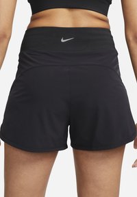 Mujer vestida con pantalones cortos deportivos negros y una parte superior deportiva negra, vista desde atrás, destacando el logo de Nike en la cinturilla.