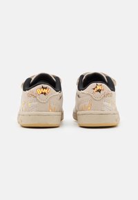 Beige sneakers med svarta detaljer, texturerad övre del i canvas, med orangea serietidningsliknande grafik "BAM!" och "POW!" på häldelen.