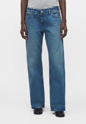 Person iført blå jeans med lige ben, grå top og sorte sko, står mod en ensfarvet lys baggrund.