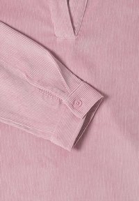 Camisa de pana rosa con escote en V y mangas largas. Presenta una textura de rayas verticales y un detalle de botón en el puño de la manga.