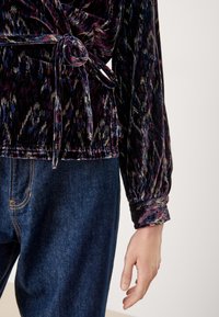 Top in velluto di un viola scuro con un motivo astratto multicolore, dotato di un design avvolgente, maniche lunghe e vita legata. Abbinato a jeans blu.