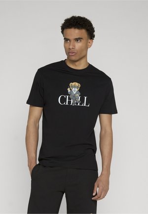 Jeune homme portant un t-shirt noir avec un graphisme d’ours en costume assis sur le mot « CHILL » sur la poitrine, sur fond clair uni.