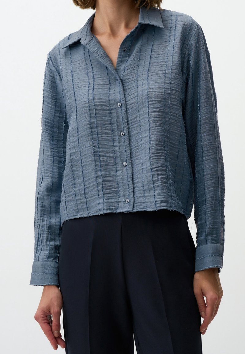 Femme portant une chemise bleue texturée à manches longues boutonnée avec col et un pantalon noir taille haute.