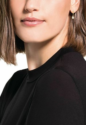 Goldene Creolen mit einem kleinen ovalen Akzent. Das Modell trägt ein schwarzes, hochgeschlossenes Oberteil, das glattes Haar und dezentes Make-up zur Geltung bringt.