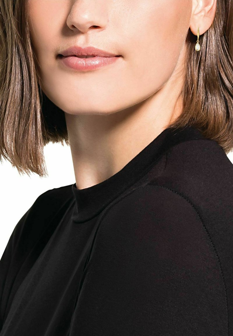Orecchino a cerchio in oro con un piccolo accento ovale. Il modello indossa un top nero a collo alto, mostrando capelli lisci e trucco neutro.