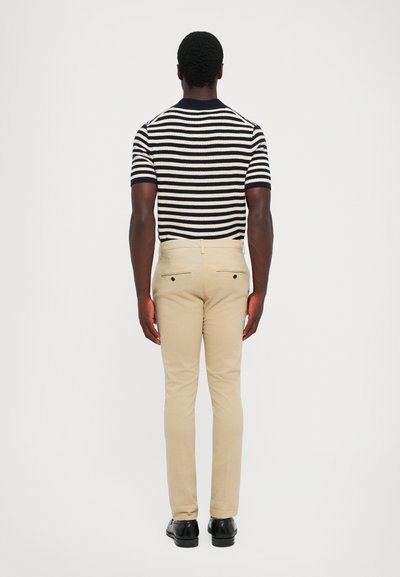Uomo in piedi di spalle, con una camicia a maniche corte a righe bianche e nere, pantaloni beige e scarpe eleganti nere, su uno sfondo uniforme.