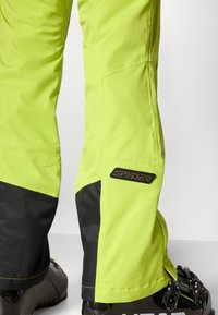 Neon gule snøbukser med svarte aksenter og logo. Laget av vanntett materiale, med en avsmalnende design og forsterkede sømmer.