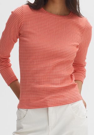 Femme portant un haut ajusté à manches longues rayé rouge et rose avec un pantalon blanc, mains dans les poches, sur un fond uni.