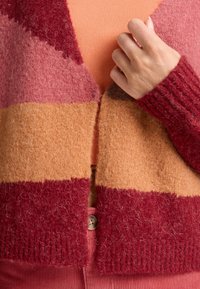 Gebreide cardigan in tinten van rood, roze en beige met een fuzzy textuur, geribde mouwen en een knoopsluiting aan de onderkant.