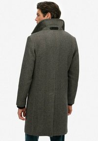 Grijze herringbone wollen jas met een hoge kraag, zwarte accenten op de manchetten en een knoop aan de achterkant, met een rechte snit en lange mouwen.