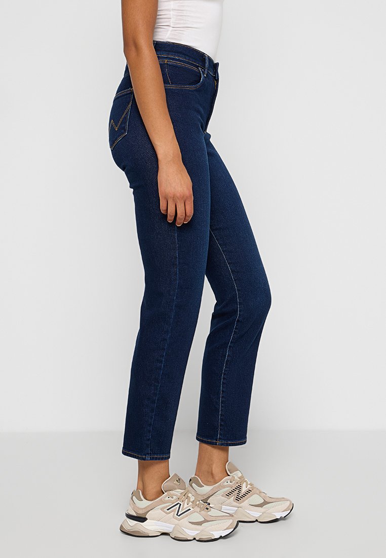 Jean en denim bleu à coupe droite, avec des coutures contrastées, deux poches arrière, et portés avec des baskets beiges. Design simple, taille moyenne.