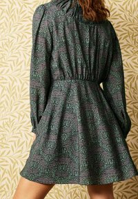 Vestido corto de manga larga en tela verde oscura con un patrón floral en verde claro y morado. Presenta un escote y una cintura fruncidos.