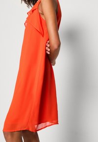 Robe orange sans manches avec des accents à volants, une coupe ample et un ourlet à hauteur du genou. Fabriquée en tissu léger, elle présente une texture lisse.