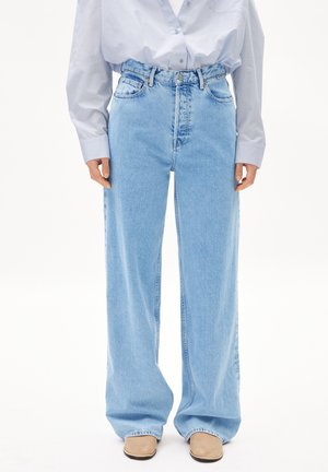 Person iført lyseblå vidde bukser med høj talje, kombineret med en løs hvid skjorte med knapper og beige sko, vist fra taljen og ned.