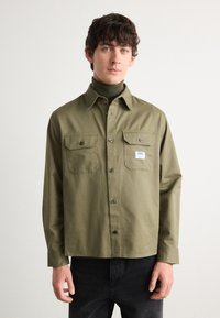 Olive green button-up shirt with two chest pockets, black buttons, and a small brand label. Long sleeves with a straight hem and relaxed fit.