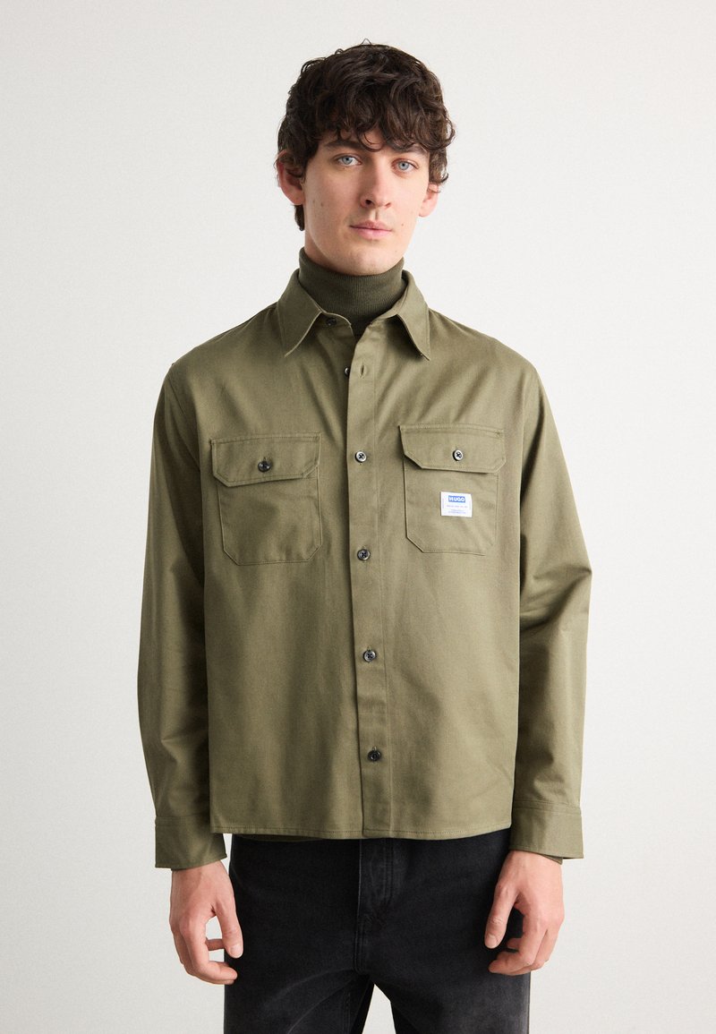 Olive green button-up shirt with two chest pockets, black buttons, and a small brand label. Long sleeves with a straight hem and relaxed fit.