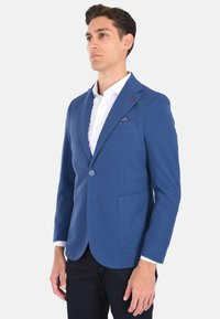 Blazer blu con texture, dotato di due tasche frontali, chiusura con un solo bottone e rever. Indossato sopra una camicia bianca, con un fazzoletto nel taschino.