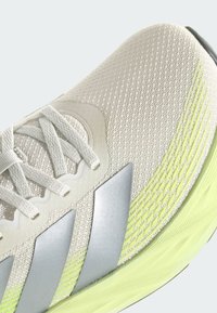 Close-up of beige mesh running shoe with silver stripes, white laces, and light green textured sole.