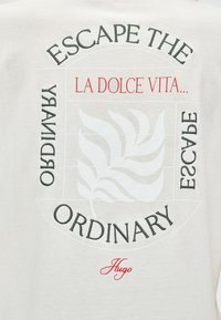 Tissu blanc avec un motif circulaire comprenant une feuille stylisée et le texte : « Escape the ordinary », « La Dolce Vita » et « Hugo » en polices rouge et noire.