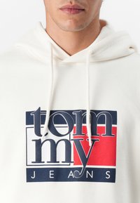 Bílá mikina s kapucí a šňůrkami s modro-červeným grafickým logem, které v tučném písmu zní "tom my JEANS". Měkká textilní struktura.