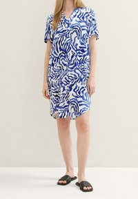 Šaty s krátkým rukávem v modro-bílé abstraktní vlnité vzoru, vyrobené z lehkého materiálu, s výstřihem do V a detaily na bočních rozparech, doplněné černými sandály.