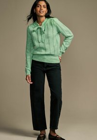Pull en maille vert menthe avec un col à nœud papillon, présentant des rayures verticales et des poignets côtelés, associé à un jean noir à jambe droite et des chaussures sombres.