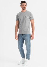Camiseta de algodón gris con escote redondo, que presenta pequeñas manchas negras y un logotipo negro circular en el lado izquierdo del pecho. Llevada con jeans de color azul claro.