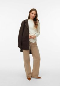 Beige wijde jeans gecombineerd met een crèmekleurige top met lange mouwen en een donkergbruine suède jas. Puntige bruine hakken maken de outfit compleet.