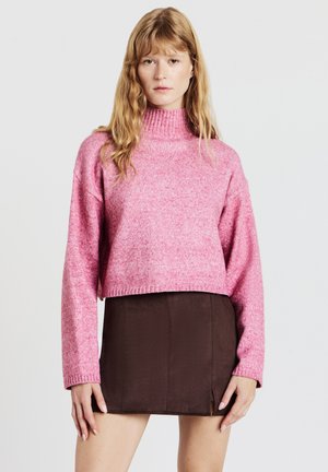 Rosa, kurz geschnittener Pullover mit hohem Kragen und gerippten Bündchen, kombiniert mit einem braunen Minirock mit seitlichem Schlitz. Glatte Textur insgesamt.