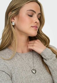 Femme aux longs cheveux blonds portant un pendentif en forme de cœur argenté et de grandes boucles d'oreilles argentées assorties sur un pull en tricot gris.
