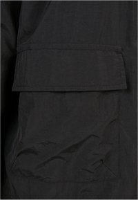 Tissu noir pour veste avec une surface texturée ; présente une grande poche à rabat cousue sur le côté inférieur gauche. Design minimaliste et fonctionnel.