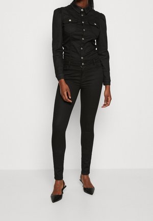 Frau trägt eine taillierte schwarze Jacke mit Knöpfen und Vordertaschen, schwarze enge Hose und schwarze Slingback-Pumps mit spitzen Zehen, steht vor einem weißen Hintergrund.