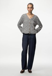 Pelēks adīts kardigans ar V-veida kaklu, ar teksturētu rakstu un brūniem pogām. Kombinēts ar tumši ziliem taisnās kājas džinsiem.
